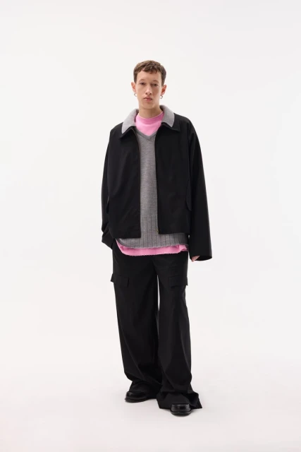 Full-length studio shot of a young adult male model wearing a black jacket, vest, pink top, and black pants against a white background.