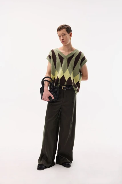 A young adult with auburn hair is wearing a sleeveless argyle sweater, olive slacks, and black loafers, holding a beaded purse indoors.