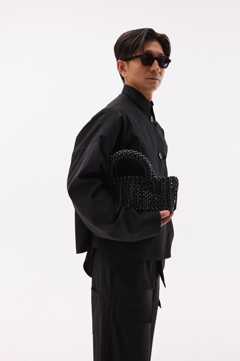 A stylish Asian man in sunglasses and a black outfit stands against a white background, holding a beaded black handbag, showcasing fashion.