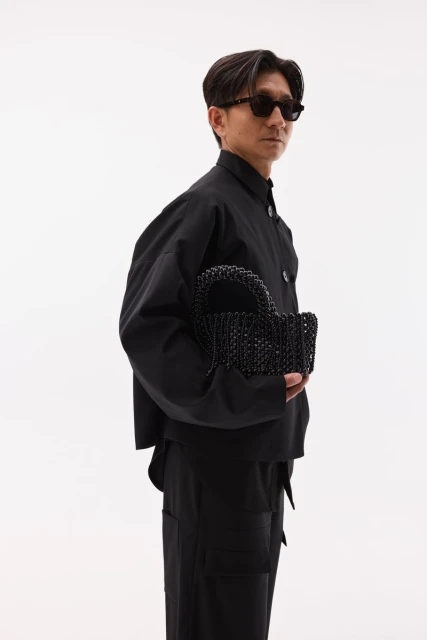 A stylish Asian man in sunglasses and a black outfit stands against a white background, holding a beaded black handbag, showcasing fashion.