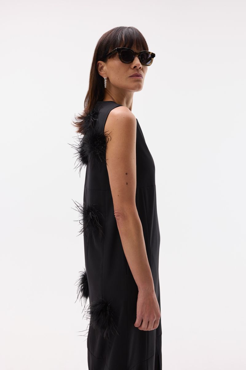 Model wearing dark sunglasses and a sleeveless black dress adorned with black feather accents along the back against a white studio sweep.