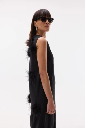 Model wearing dark sunglasses and a sleeveless black dress adorned with black feather accents along the back against a white studio sweep.
