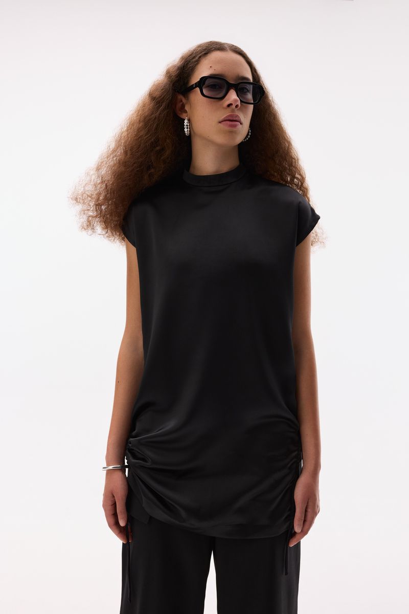 Fashion portrait of a woman with curly hair wearing black top and pants in a minimalist studio setting with soft, even lighting.