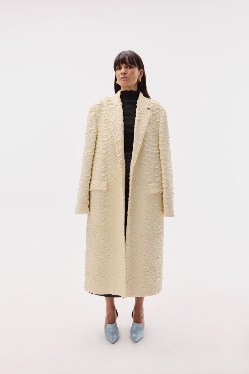 A woman in an off-white textured coat, black top, and blue heels stands in a full shot against a bright white backdrop in a fashion setting.