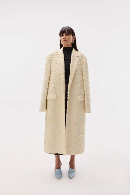 A woman in an off-white textured coat, black top, and blue heels stands in a full shot against a bright white backdrop in a fashion setting.