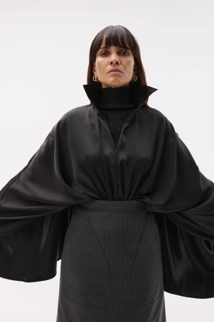 A woman poses indoors, wearing a black top with a tall collar and flared sleeves, a gray skirt, and gold hoop earrings against a white backdrop.