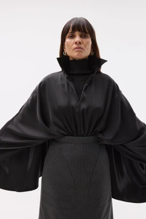 A woman poses indoors, wearing a black top with a tall collar and flared sleeves, a gray skirt, and gold hoop earrings against a white backdrop.
