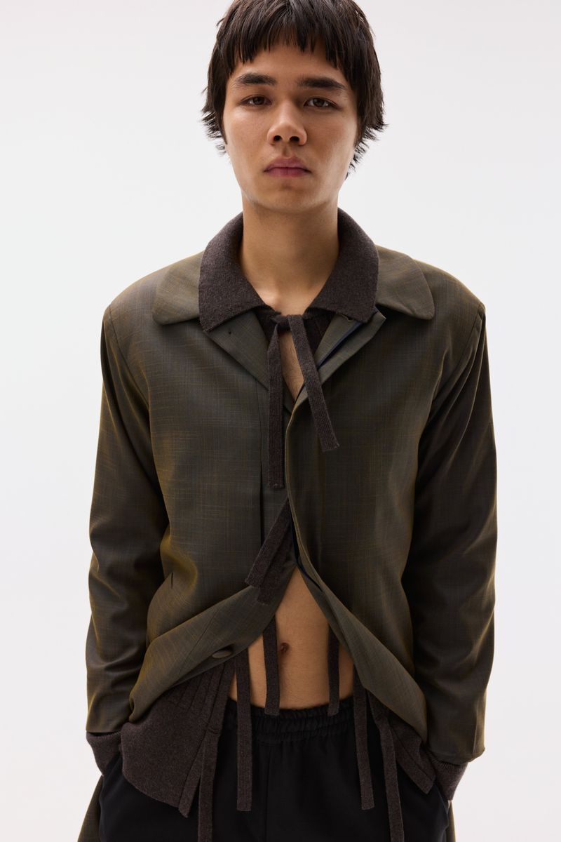 Model wearing an open olive green tailored jacket with hanging ribbon details over a dark knit against a plain white background.