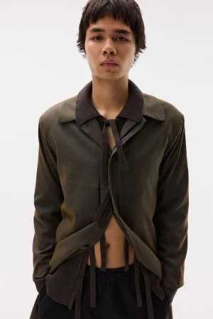 Model wearing an open olive green tailored jacket with hanging ribbon details over a dark knit against a plain white background.