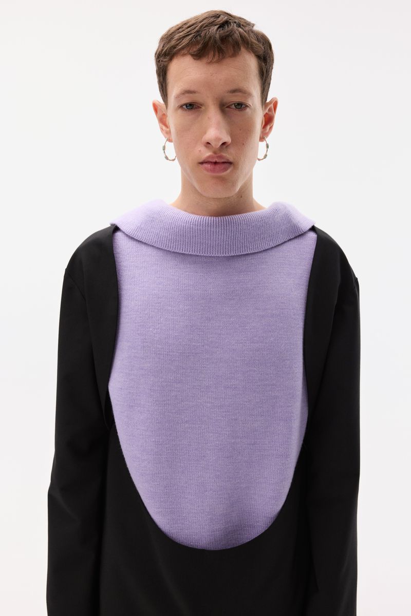 Model wearing a lavender knit panel top with black sleeves and silver hoop earrings against a white studio background.