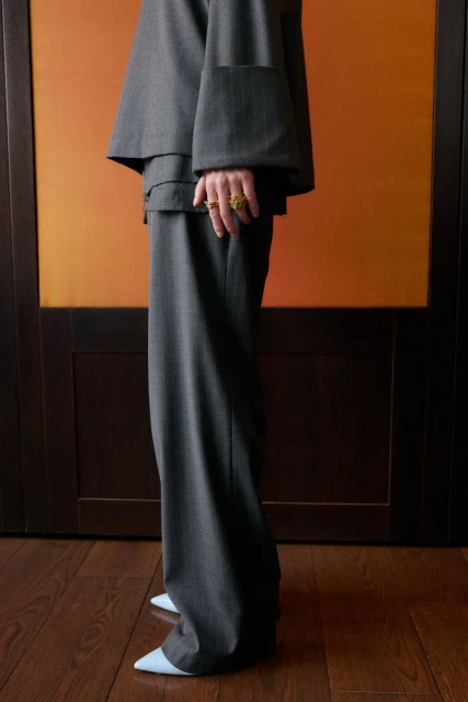 Fashion shot of a person wearing a gray, oversized pantsuit with wide sleeves, paired with pale blue heels, against an orange background.
