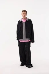 Model standing against a white seamless background wearing a black jacket, grey knit sweater, pink underlayer, and wide-leg black pants.