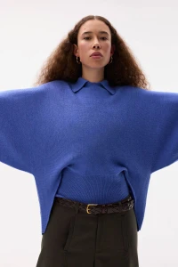 Model wearing a wide-sleeved blue collared sweater and dark trousers against a white backdrop