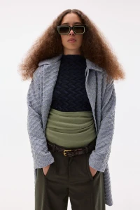 Model wearing dark sunglasses, textured grey coat, patterned top and dark trousers against a white background