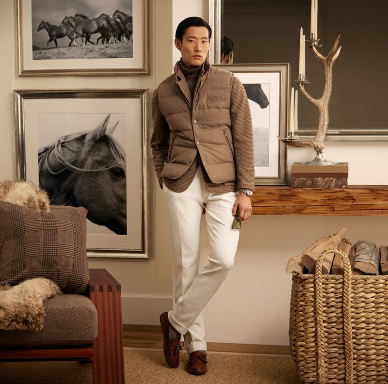 Stylish Asian man in a puffer vest and white pants stands indoors with horse art and classic decor, showcasing fashion and interior design.