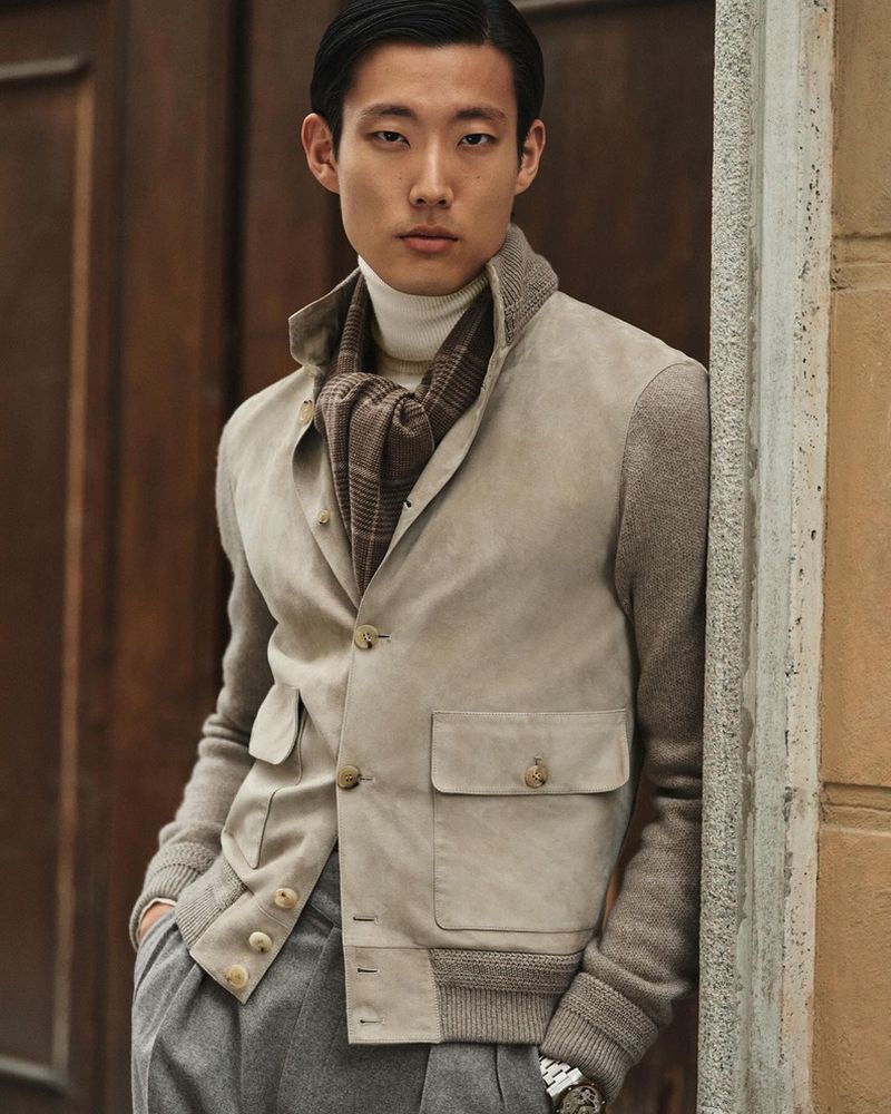 Portrait of a young Asian man in a stylish, casual outfit posing outdoors, featuring a beige jacket, turtleneck, grey pants, and scarf.