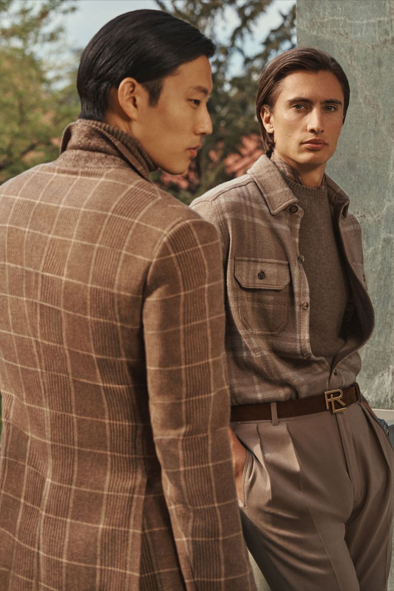 Two men pose outdoors in brown and beige toned jackets, button-down shirt, sweater, pants and a belt with logo in a medium shot.