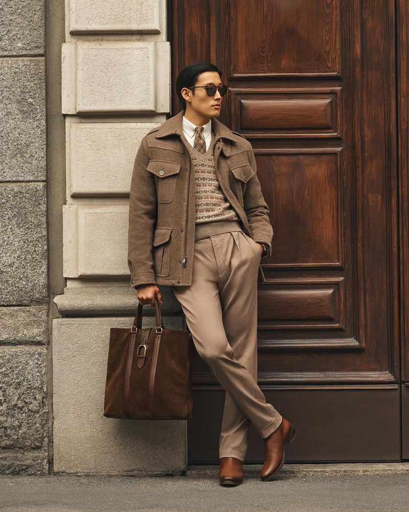 An Asian man leans against a door outdoors wearing a brown jacket, patterned sweater, brown slacks, sunglasses, and holding a brown leather tote.