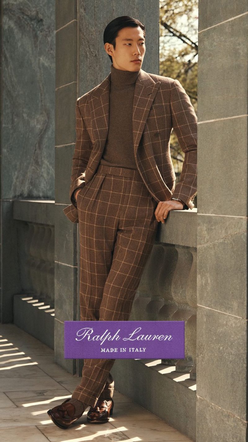 An Asian male model poses in a brown, checkered suit with a matching turtleneck indoors, leaning against a stone pillar.