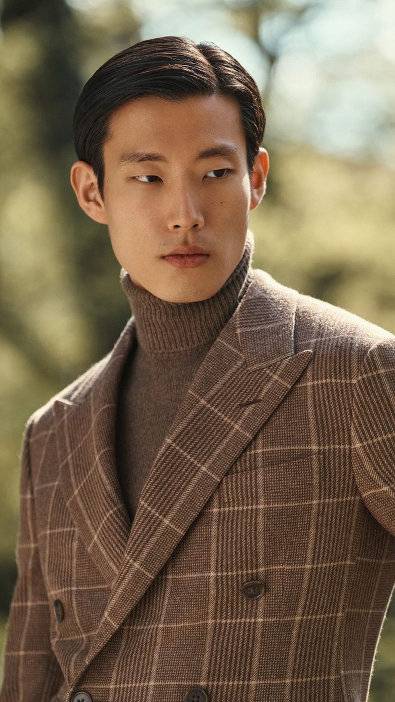Portrait of an Asian male model in a brown plaid double-breasted blazer and tan turtleneck, posing outdoors in a blurred green background.