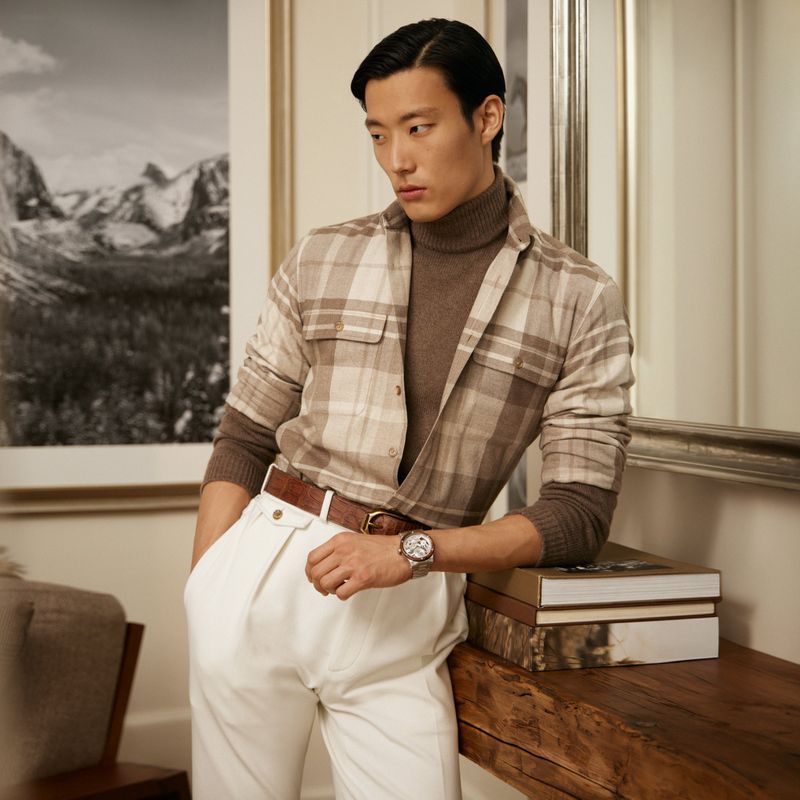 Asian man poses indoors wearing brown turtleneck, plaid shirt, and white pants. Books and framed mountain art are visible in the background.