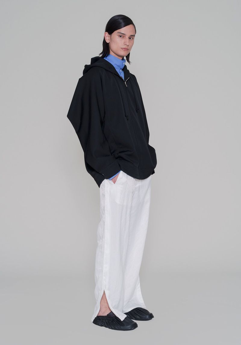 A slender person in a black hooded jacket and white trousers strikes a full-length pose against a neutral backdrop in a fashion editorial.