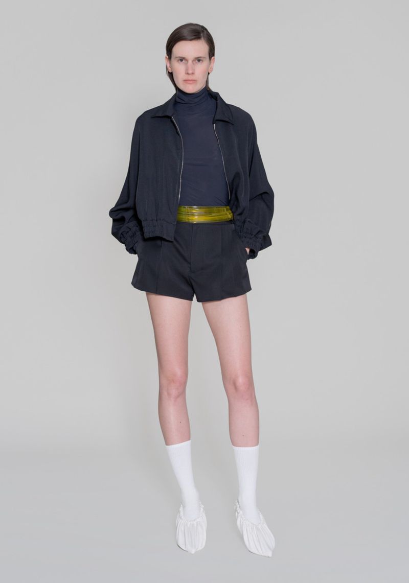 Full-length shot of a slender woman in a dark blue turtleneck, black shorts, a matching jacket, white socks, and unique white shoes.
