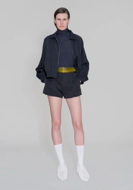 Full-length shot of a slender woman in a dark blue turtleneck, black shorts, a matching jacket, white socks, and unique white shoes.