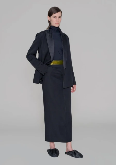 A full-length shot presents a model in a black blazer, high-neck top, and long skirt, cinched with a gold belt, set against a gray backdrop.
