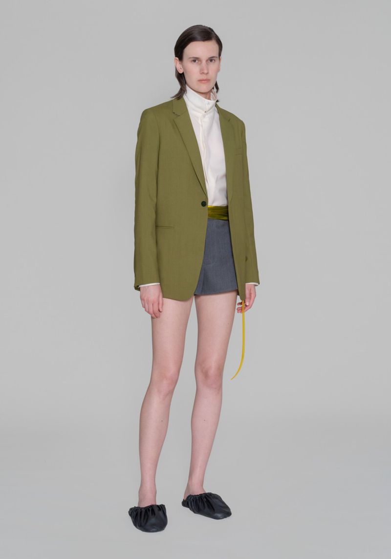 A slender fashion model with short dark hair poses in an olive blazer, white top, and gray shorts in a full shot against a gray backdrop.
