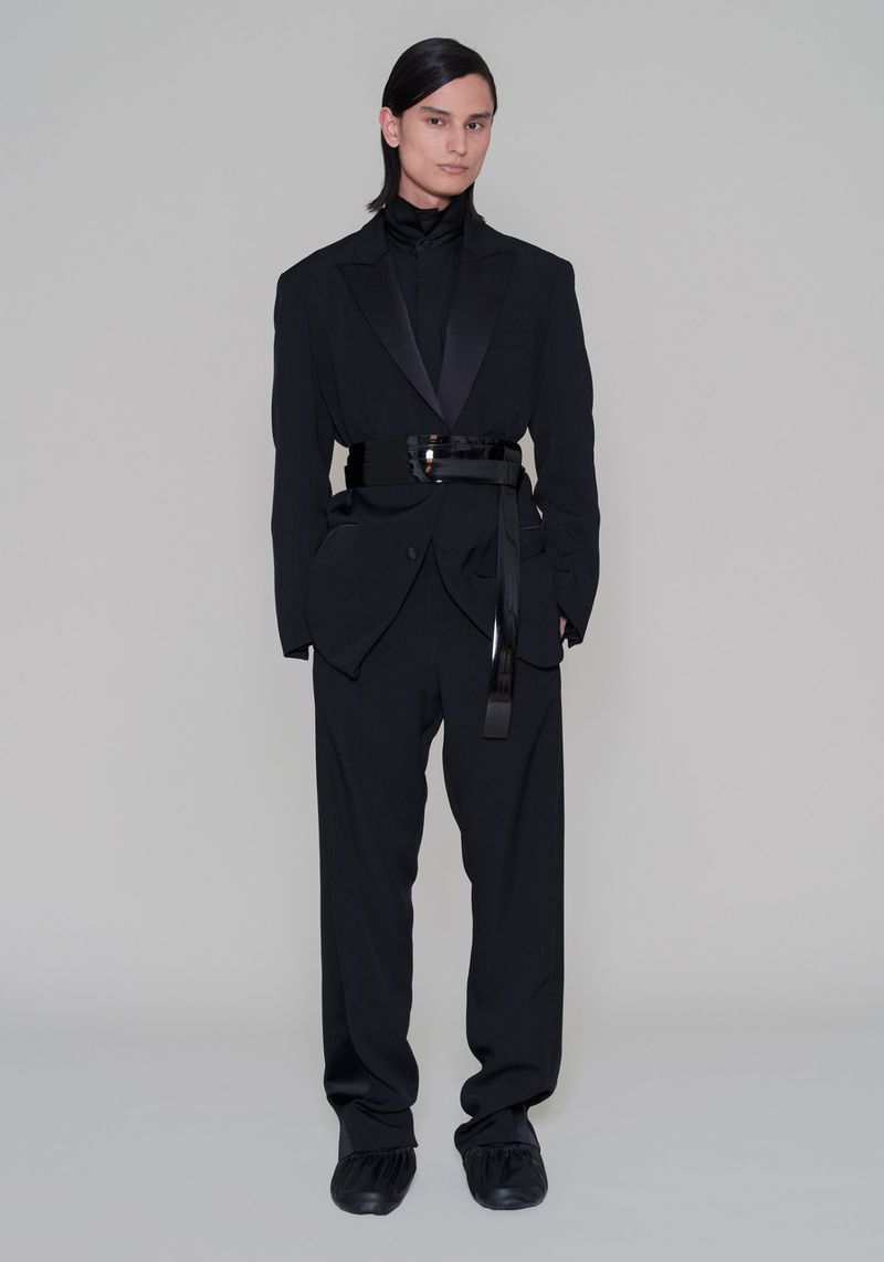 Full-length studio shot of androgynous fashion model in a black tuxedo-inspired suit with wide belt and buckled sash against a light gray background.
