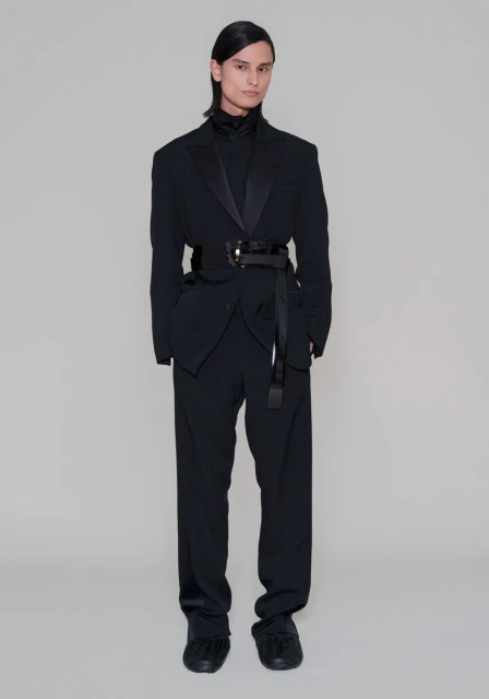 Full-length studio shot of androgynous fashion model in a black tuxedo-inspired suit with wide belt and buckled sash against a light gray background.