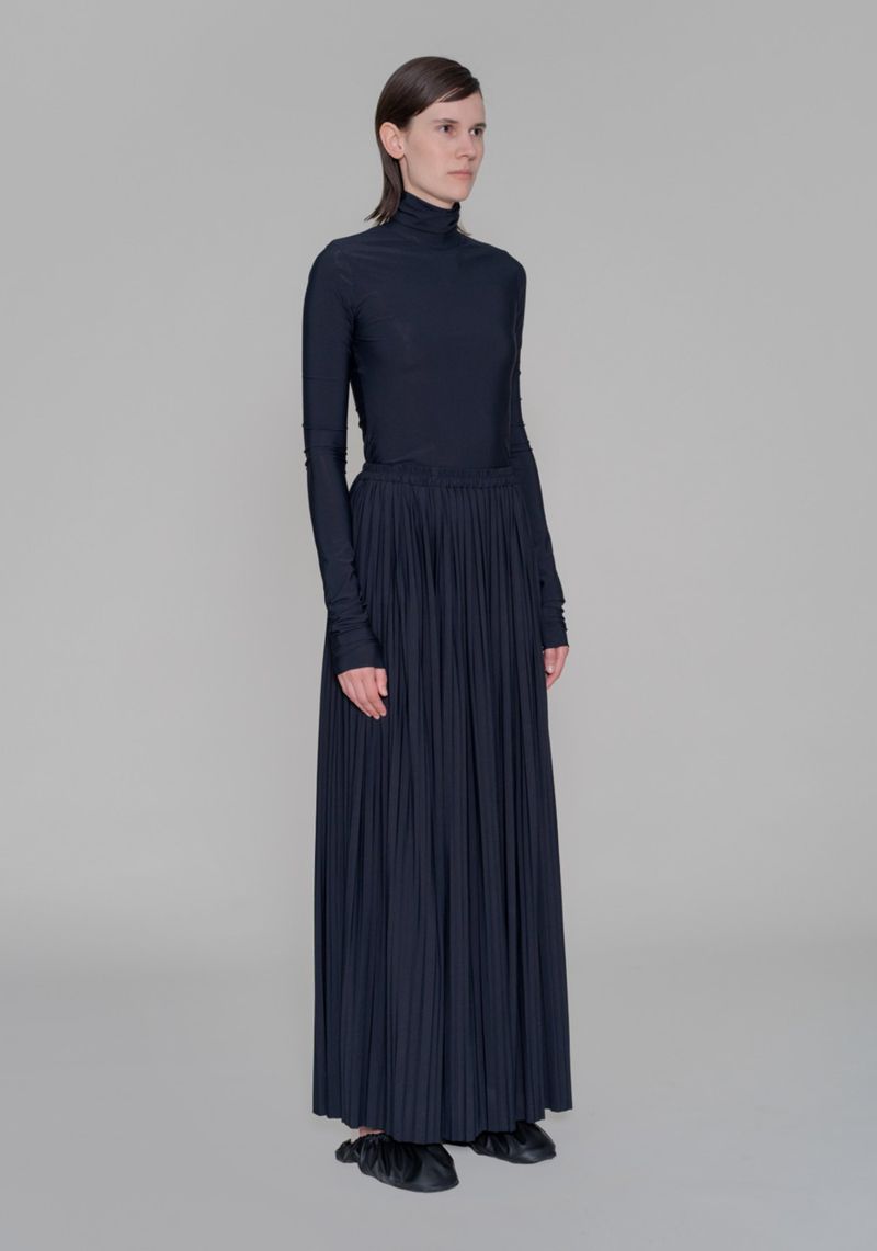 Model Jamily Wernke Meurer wearing a navy turtleneck and pleated maxi skirt against a grey backdrop.