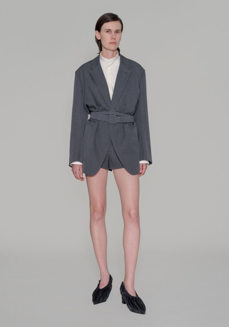 Fashion photo of a woman in a belted grey blazer and dark, sculptural heels in a studio setting, showcasing a modern, chic style.