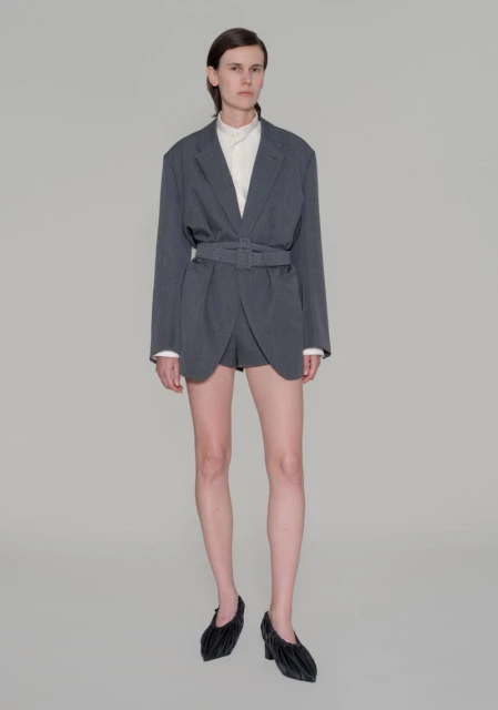 Fashion photo of a woman in a belted grey blazer and dark, sculptural heels in a studio setting, showcasing a modern, chic style.