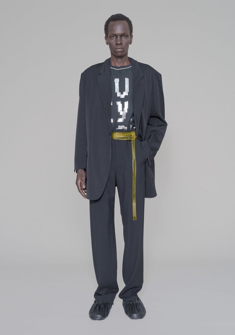 Model Marwang Lual wears an oversized dark suit with a long translucent yellow belt against a seamless grey backdrop.
