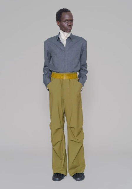 An adult male model is standing in a photo studio wearing a gray shirt, light yellow belt, and loose fitting olive-green pants looking off to the side.