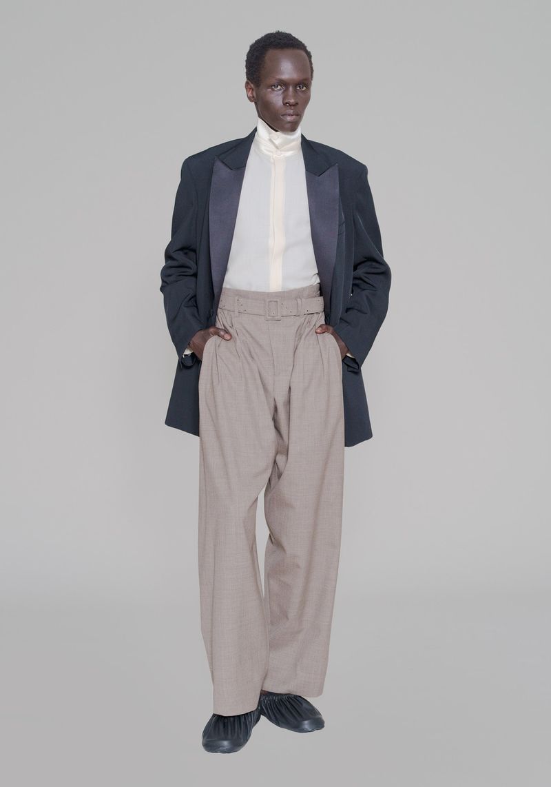Front view of a Black male fashion model in a black blazer with satin lapels, light shirt, wide tan pants, and black footwear, set against grey.