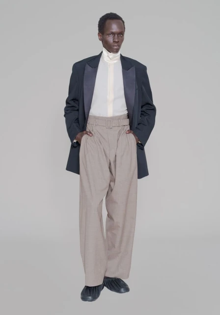 Front view of a Black male fashion model in a black blazer with satin lapels, light shirt, wide tan pants, and black footwear, set against grey.