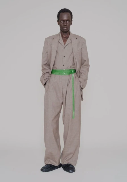 Adult male in a photo studio, wearing a gray suit with a green belt, posing against a neutral backdrop in a full shot.