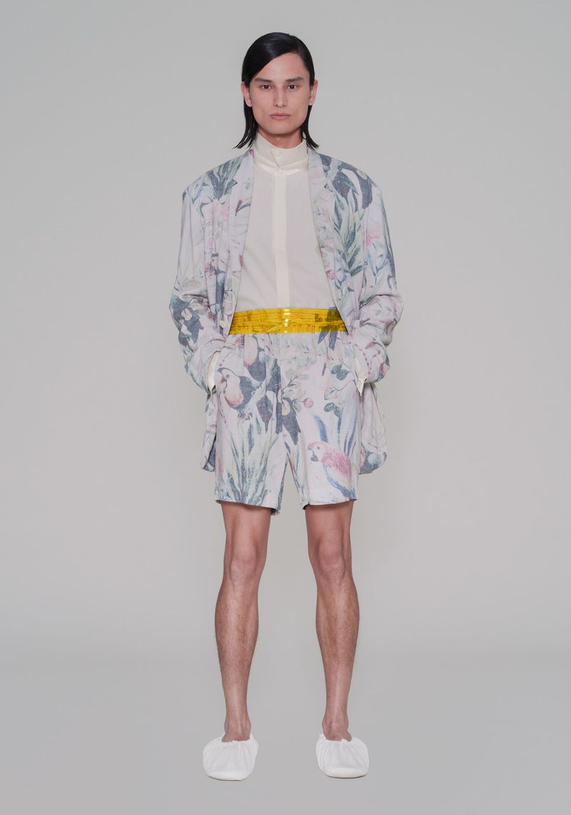 Full-length portrait of an adult posing in patterned blazer, shorts, and white slippers against a neutral studio backdrop.