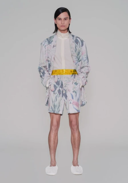 Full-length portrait of an adult posing in patterned blazer, shorts, and white slippers against a neutral studio backdrop.
