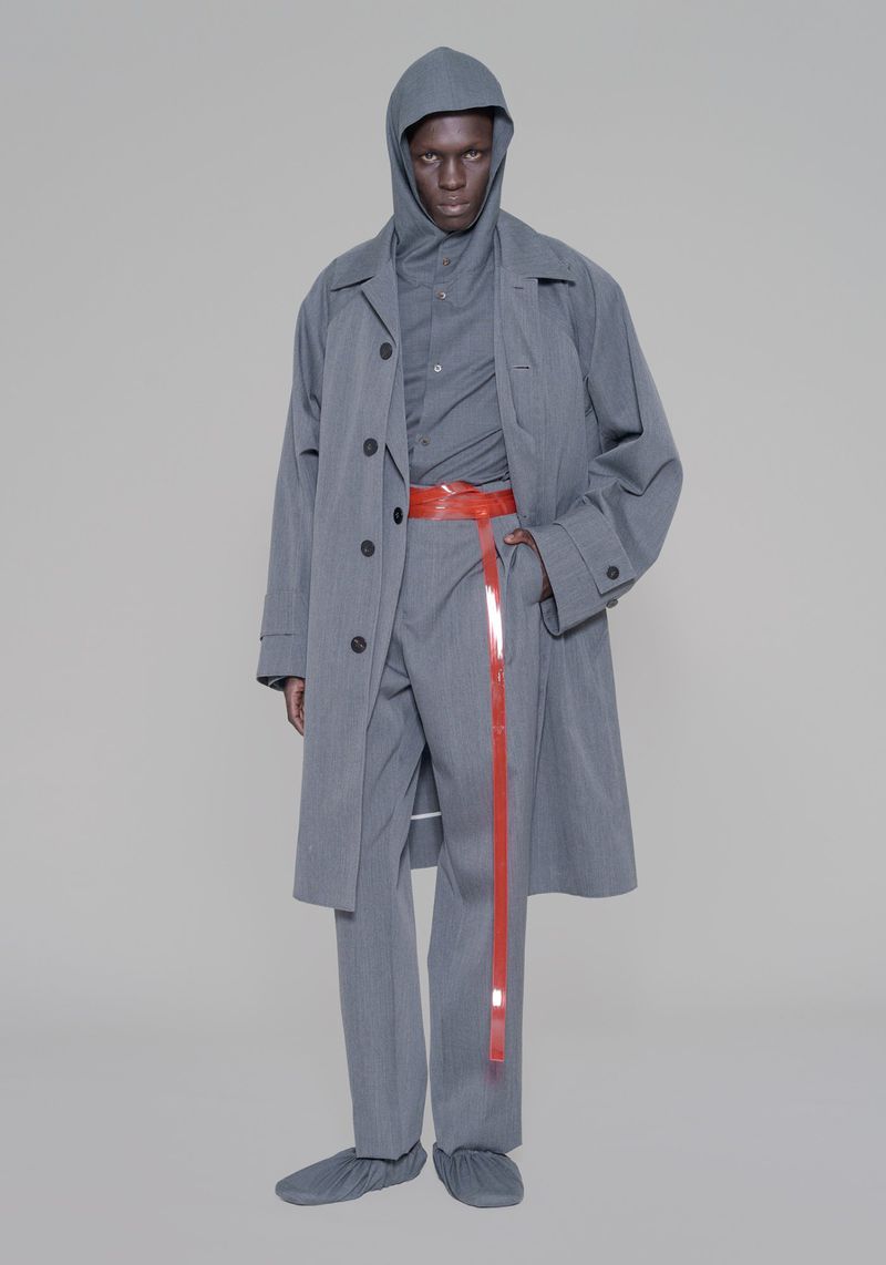 Man in grey hooded outfit and overcoat, accessorized with a long red belt against a neutral backdrop in a fashion setting.