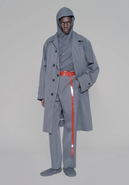 Man in grey hooded outfit and overcoat, accessorized with a long red belt against a neutral backdrop in a fashion setting.