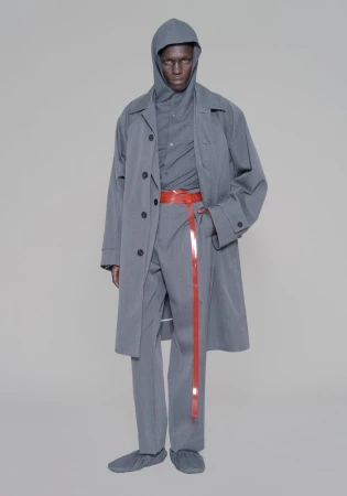Man in grey hooded outfit and overcoat, accessorized with a long red belt against a neutral backdrop in a fashion setting.