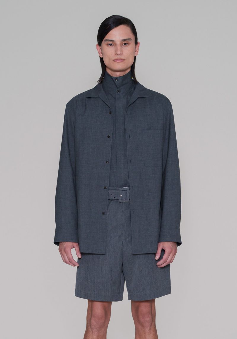 Male model in a grey collared shirt, jacket, and belted shorts poses in a studio. Full shot. Fashion editorial. Neutral backdrop.