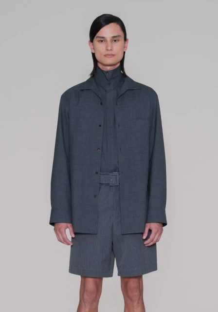 Male model in a grey collared shirt, jacket, and belted shorts poses in a studio. Full shot. Fashion editorial. Neutral backdrop.