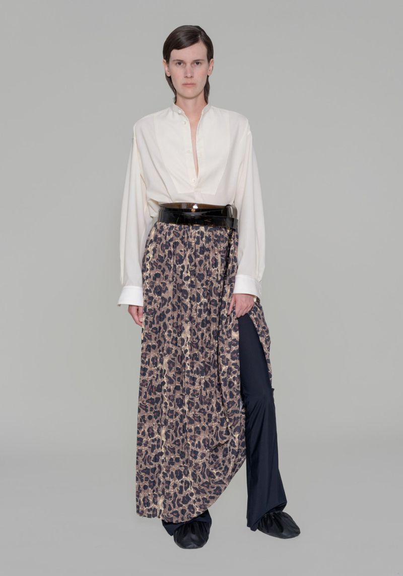 Full shot of adult female fashion model wearing a loose white button-down shirt, wide black belt, and leopard print skirt over pants in studio.