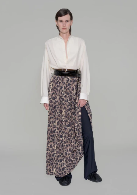Full shot of adult female fashion model wearing a loose white button-down shirt, wide black belt, and leopard print skirt over pants in studio.