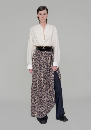 Full shot of adult female fashion model wearing a loose white button-down shirt, wide black belt, and leopard print skirt over pants in studio.